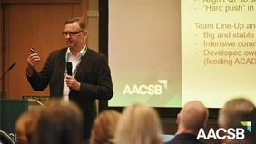 A man in a suit is standing in front of an audience, holding a microphone and a remote control, with excerpts from a presentation visible behind him.