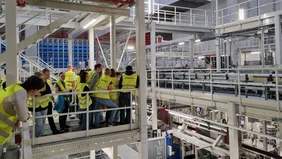 A group of people wearing yellow high-visibility vests stand on a landing and look out into the vastness of the logistics center. 