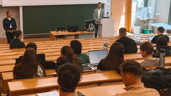 Im Hörsaal, man sieht von hinten die Studierenden die nach vorne blicken, wo zwei Referenten einen Vortrag halten.