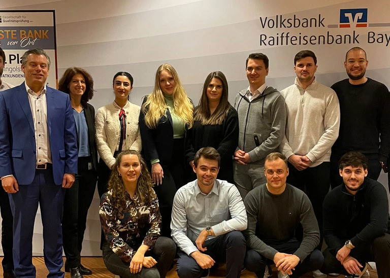 A group photo with 14 people, with the VR-Bank Bayern Mitte logo in the background.