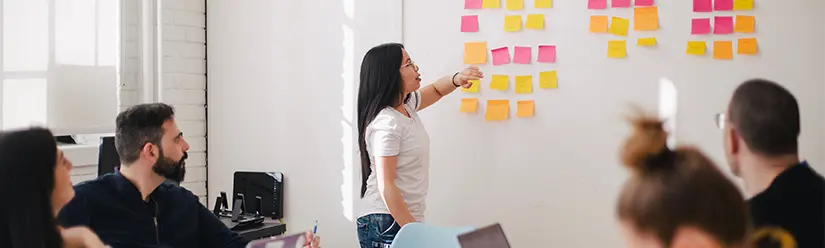 Eine Studentin steht vor ihren Kommilitonen an einer mit Post-its beklebten Tafel 