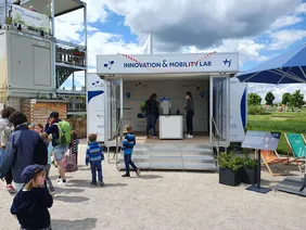 Bild zeigt das Innovation&Mobility Lab von außen mit vorbeigehenden Besuchern und einen mit Wolken bewegten Himmel.