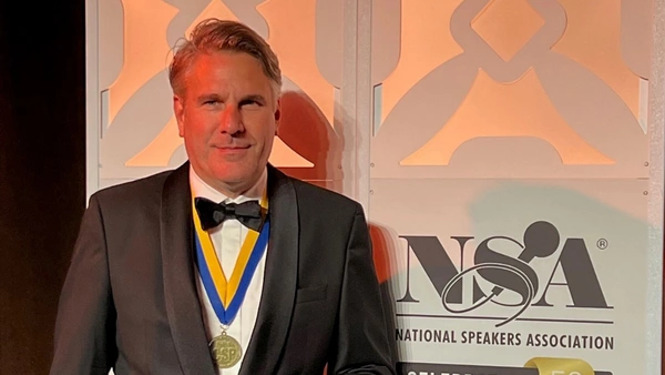 A man in black jacket with bow tie and medal in front of an illuminated wall.