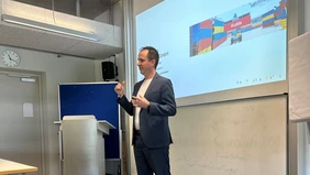 A man dressed in a dark suit stands at the front of the lecture hall, a presentation lights up behind him 